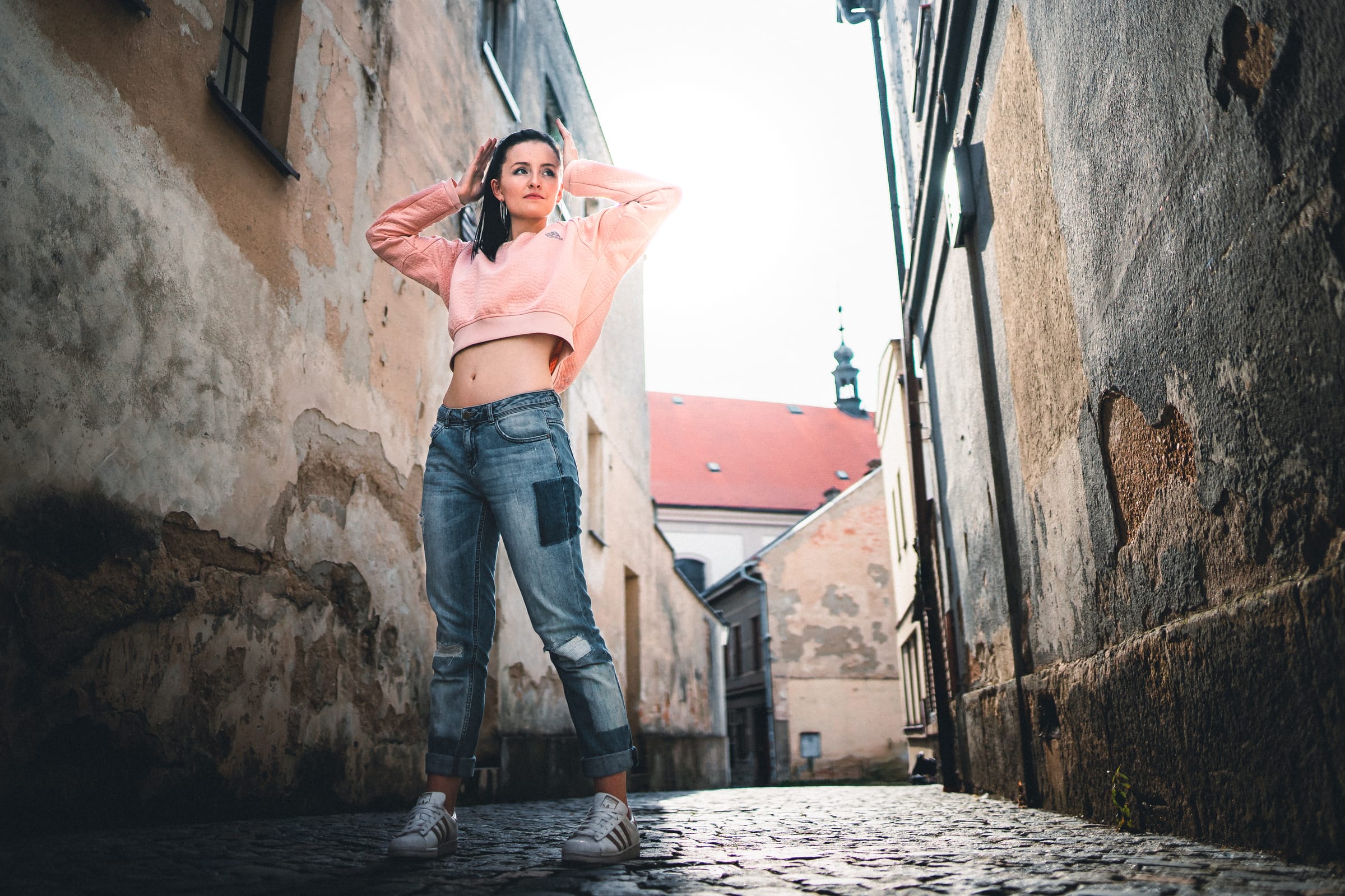 Dívka v pudrovém crop topu a džínách pózuje v úzké historické uličce, foceno z podhledu od dlažebních kostek