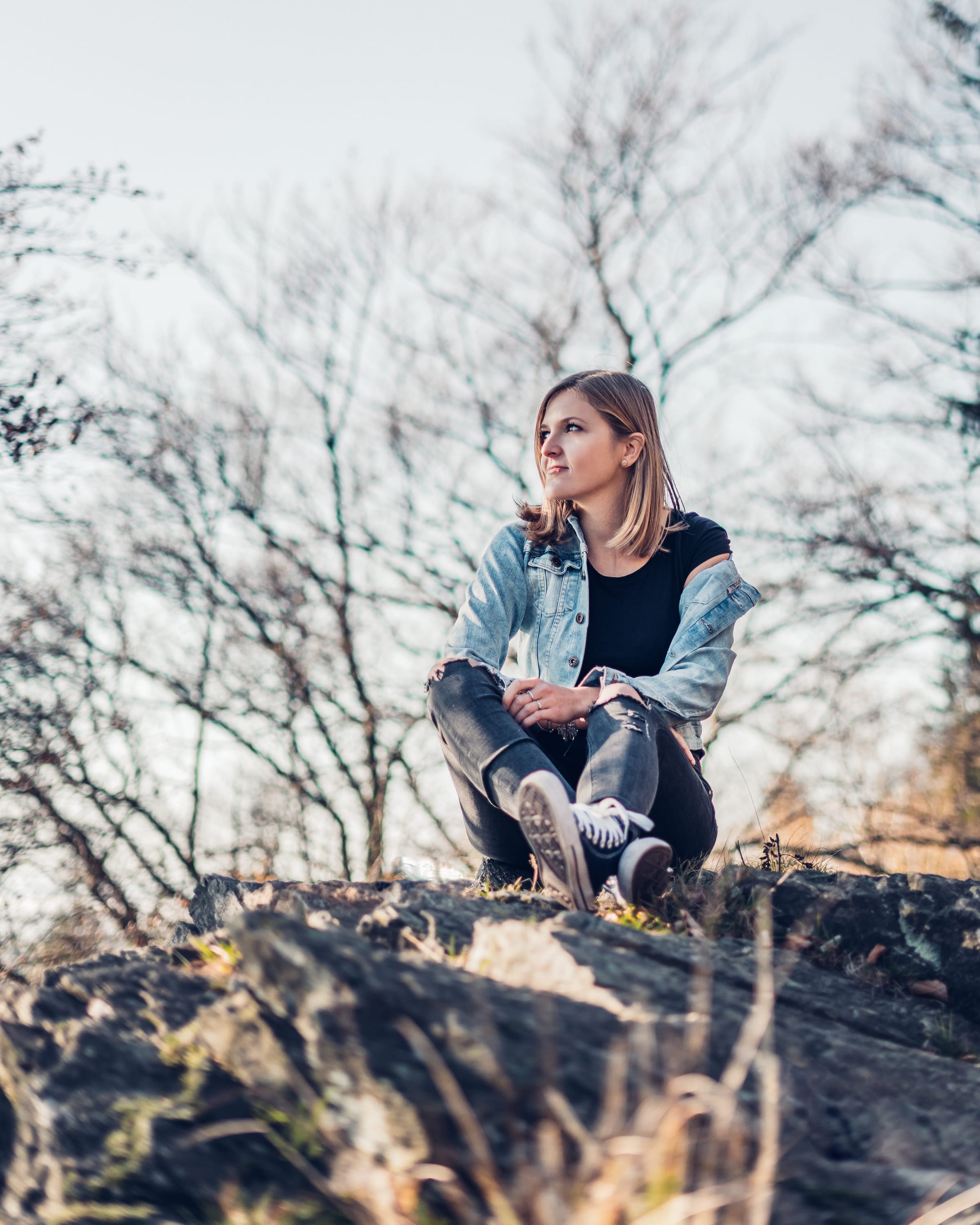 Dívka v džínové bundě sedí na skále mezi holými stromy v podzimním protisvětle