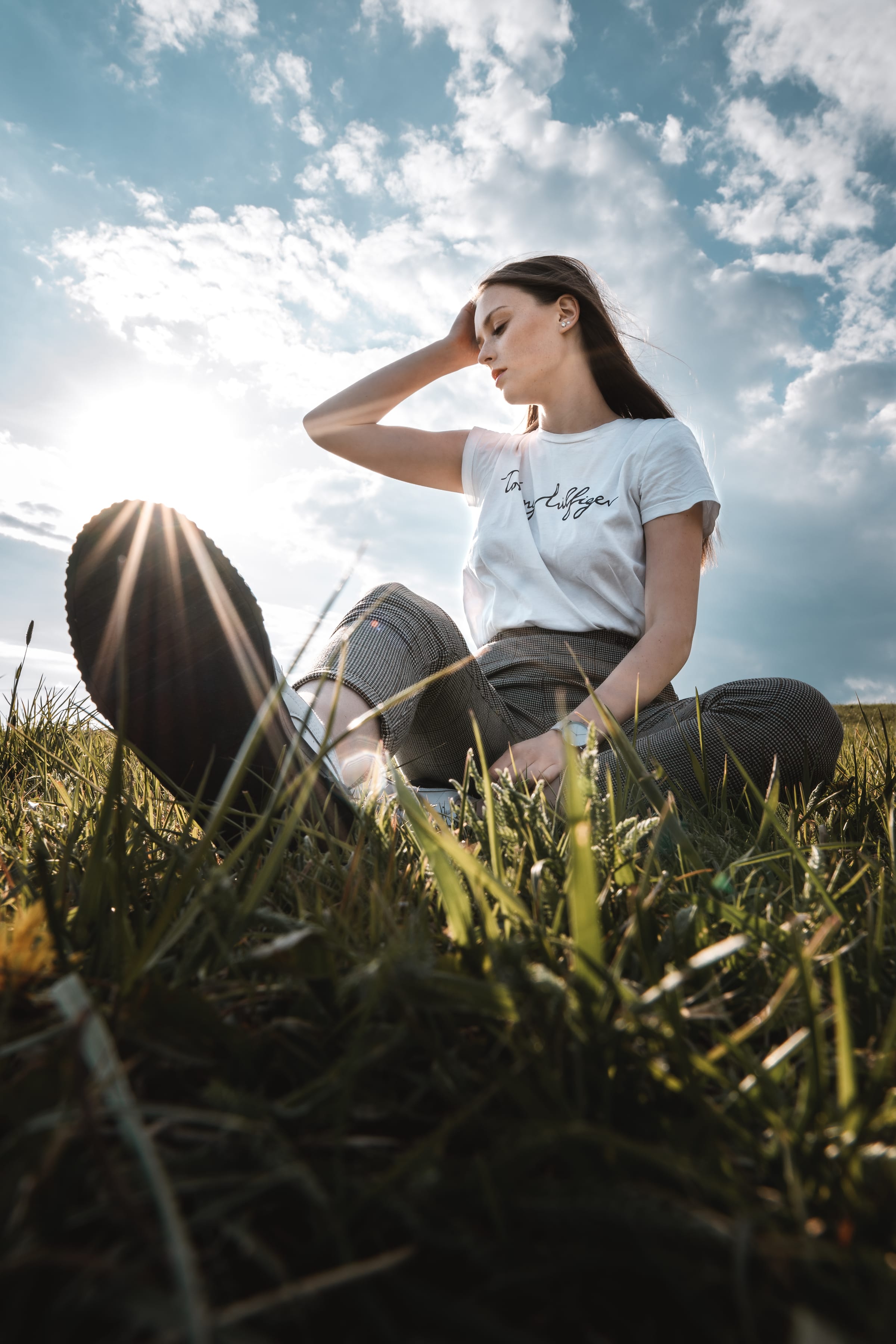 Dívka sedí v trávě proti slunci, foceno z podhledu mezi stébly pod kupovitými mraky