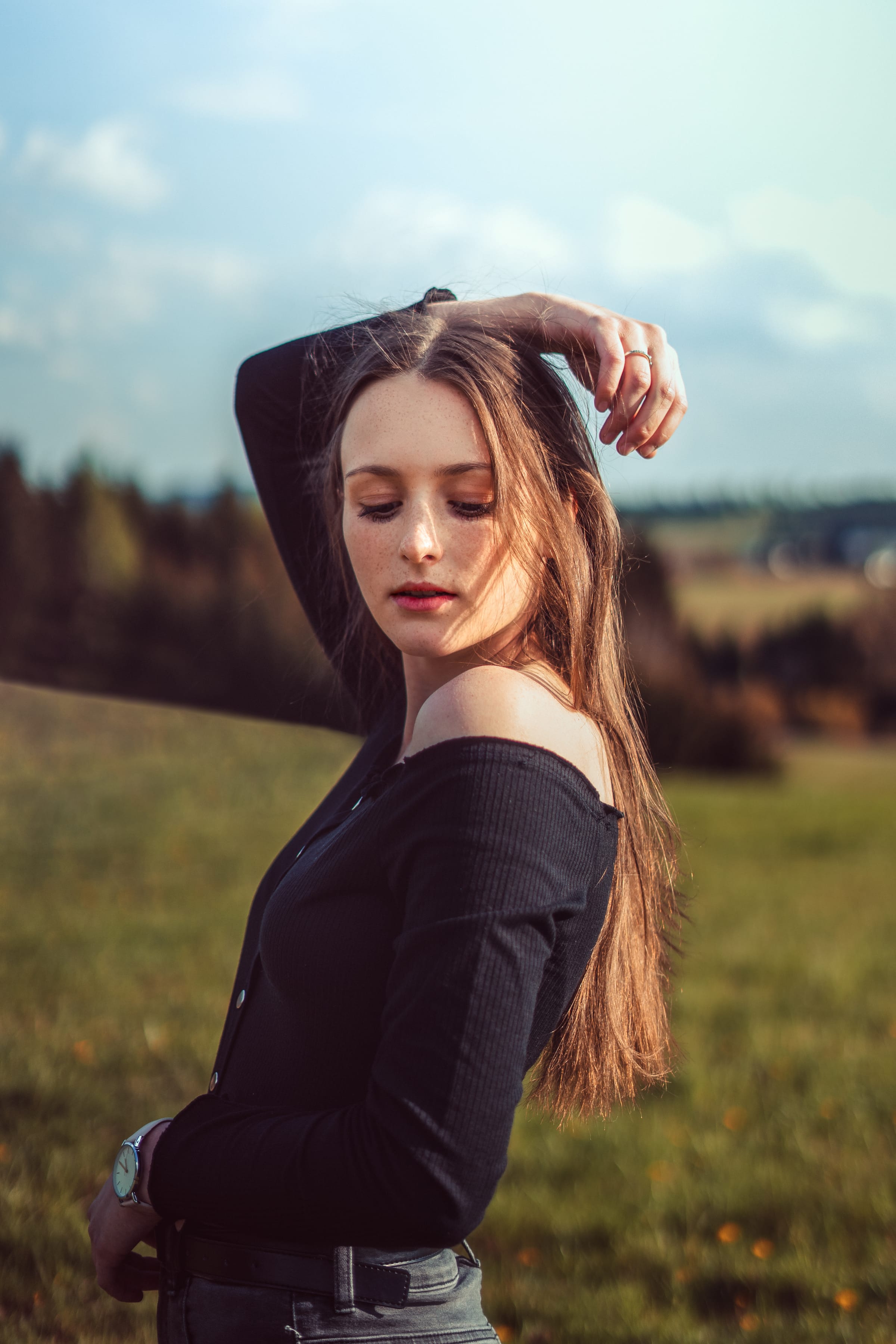 Dívka s pihami v černém topu s odhaleným ramenem na louce, ruka ve vlasech, večerní slunce