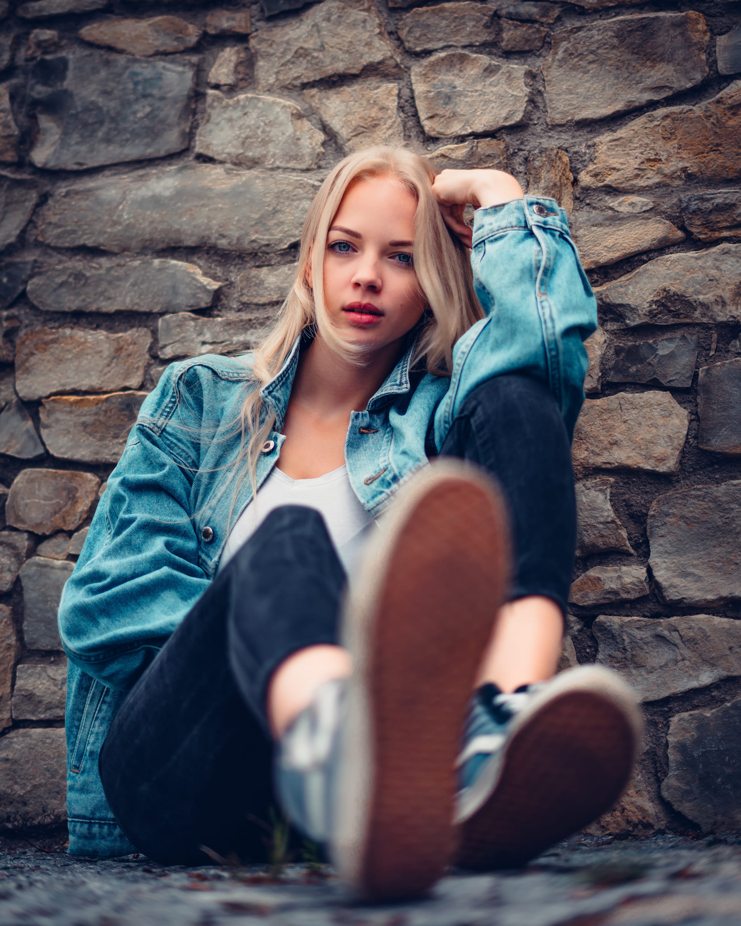 Blondýna v džínové bundě sedí opřená o kamennou zeď s nohama nataženýma k objektivu
