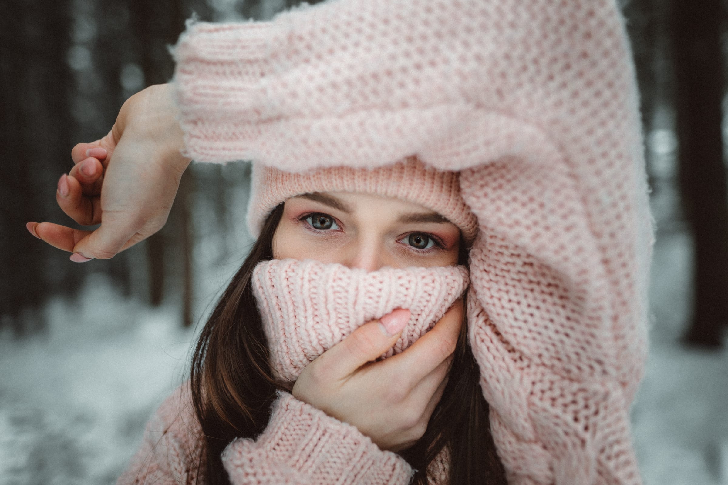 Dívka schovaná v růžovém pleteném svetru v zasněženém lese, vidět jsou jen její oči