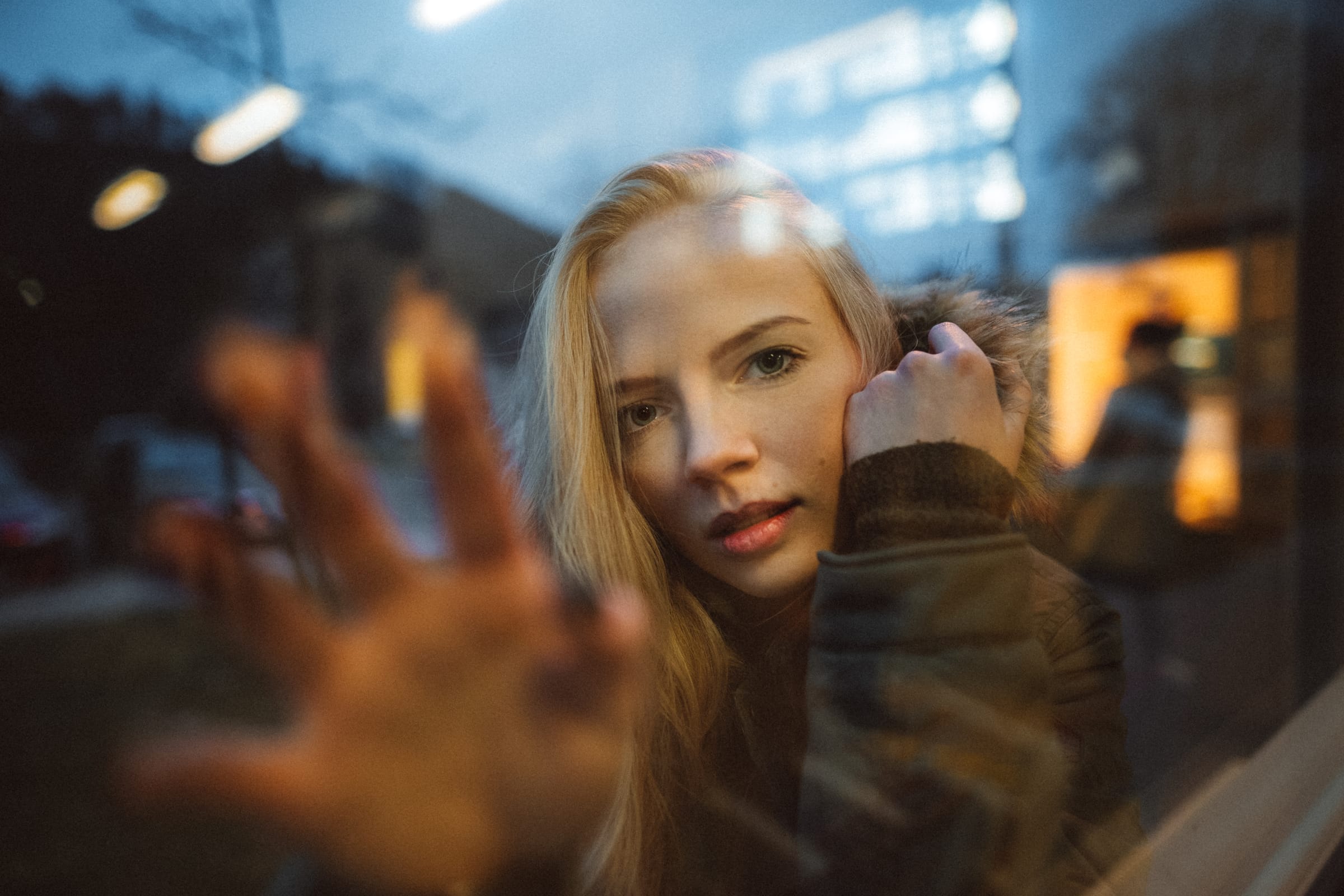 Blondýna za sklem s dlaní přitisknutou na tabuli, kolem se v okně odrážejí večerní světla města