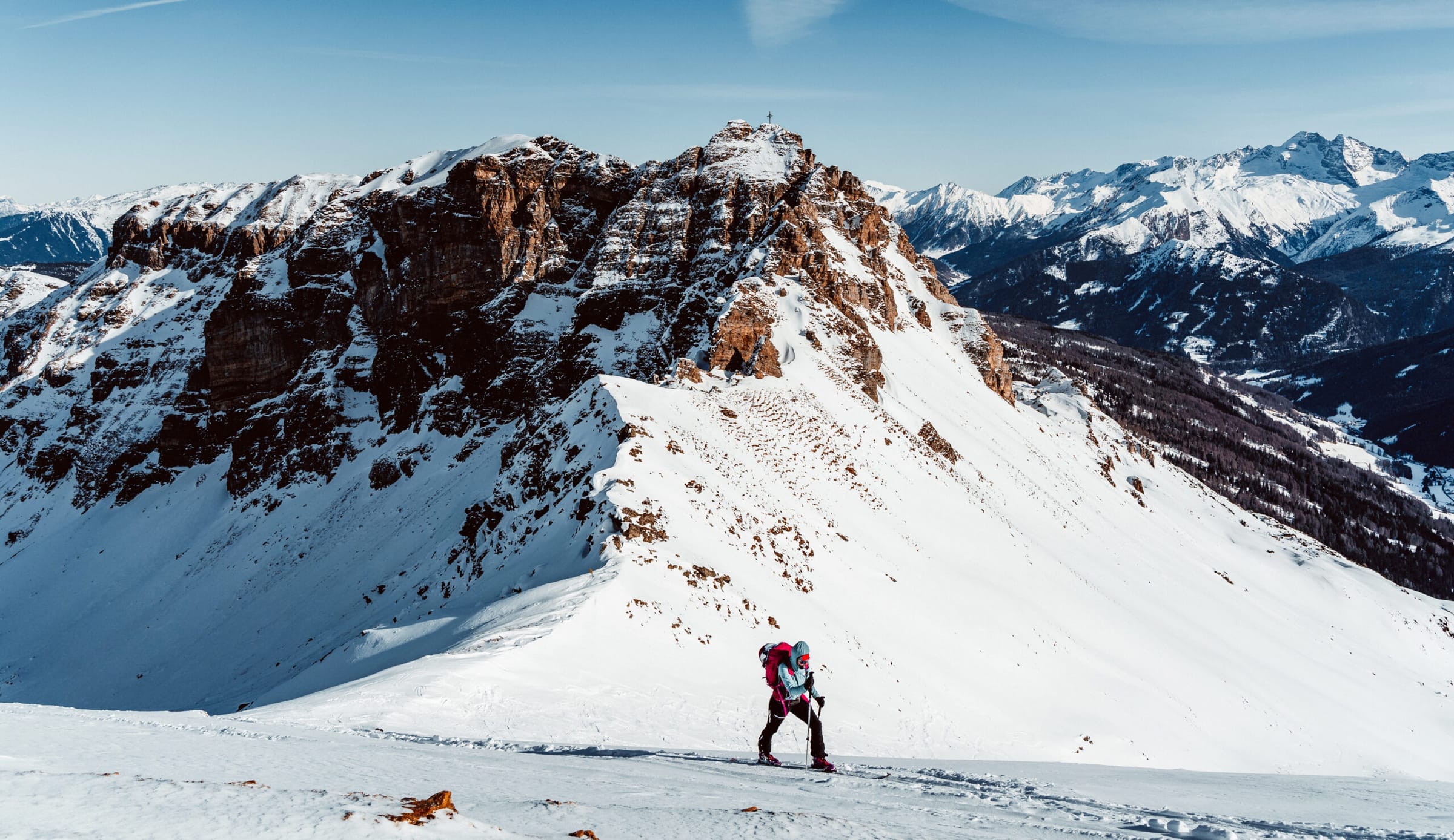 Skialpinistka s růžovým batohem stoupá na lyžích pod skalnatý vrchol s vrcholovým křížem, kolem panorama zasněžených Alp