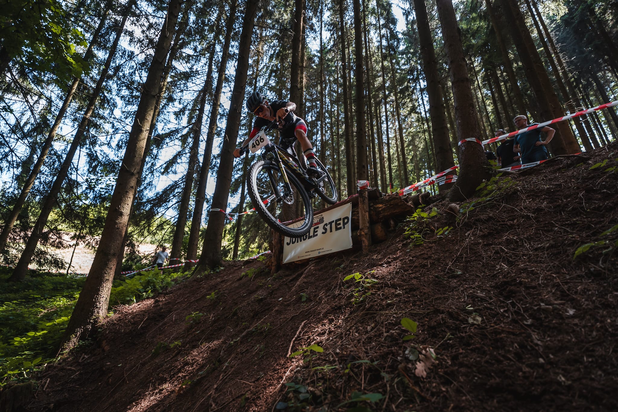Cyklista s číslem 406 sjíždí prudký lesní sráz přes dřevěnou překážku Jungle Step