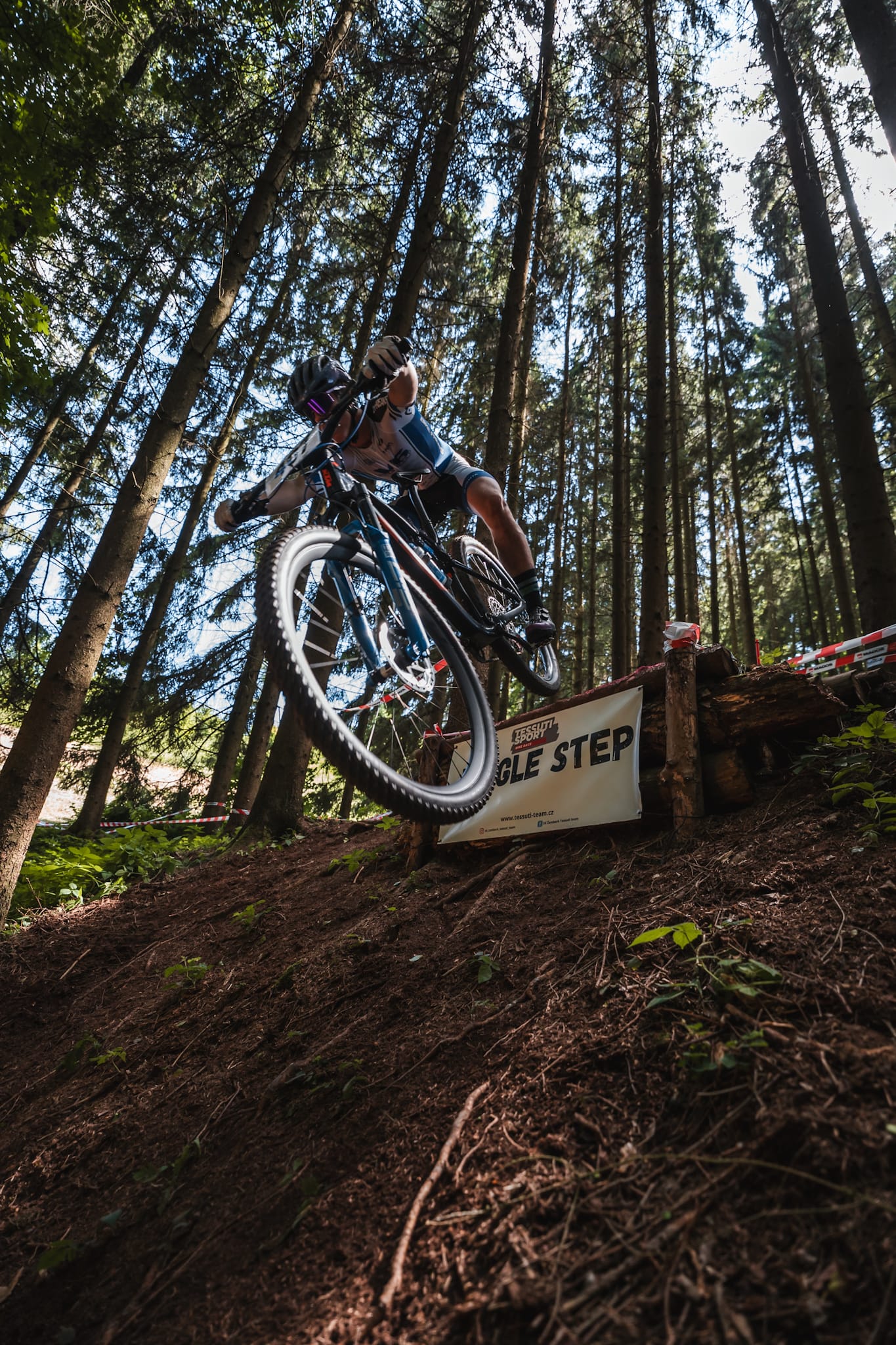 Cyklista v modrobílém dresu skáče z dřevěné překážky Jungle Step ve smrkovém lese