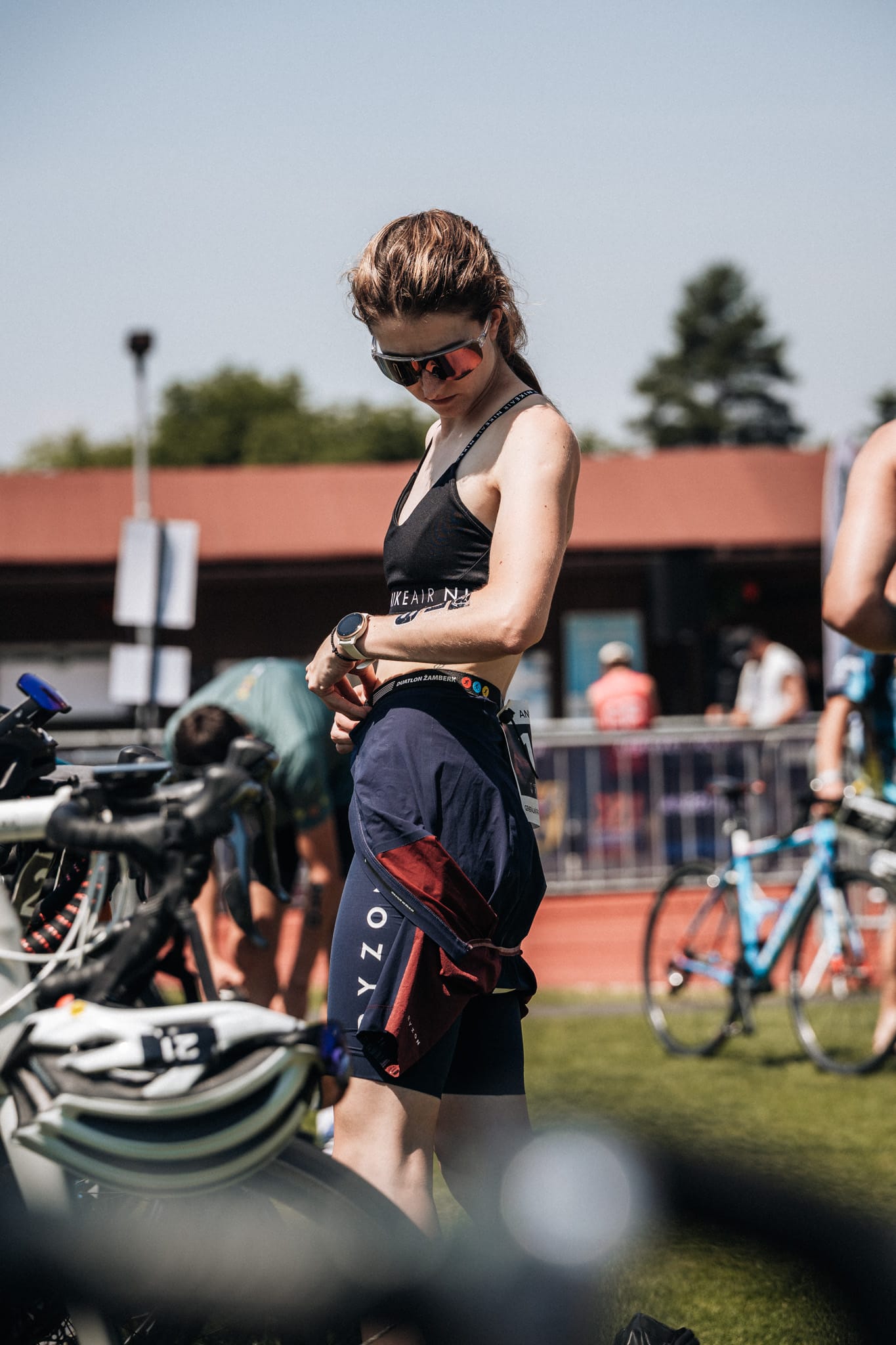 Závodnice si v depu obléká cyklistický dres přes triatlonový úbor a kontroluje hodinky