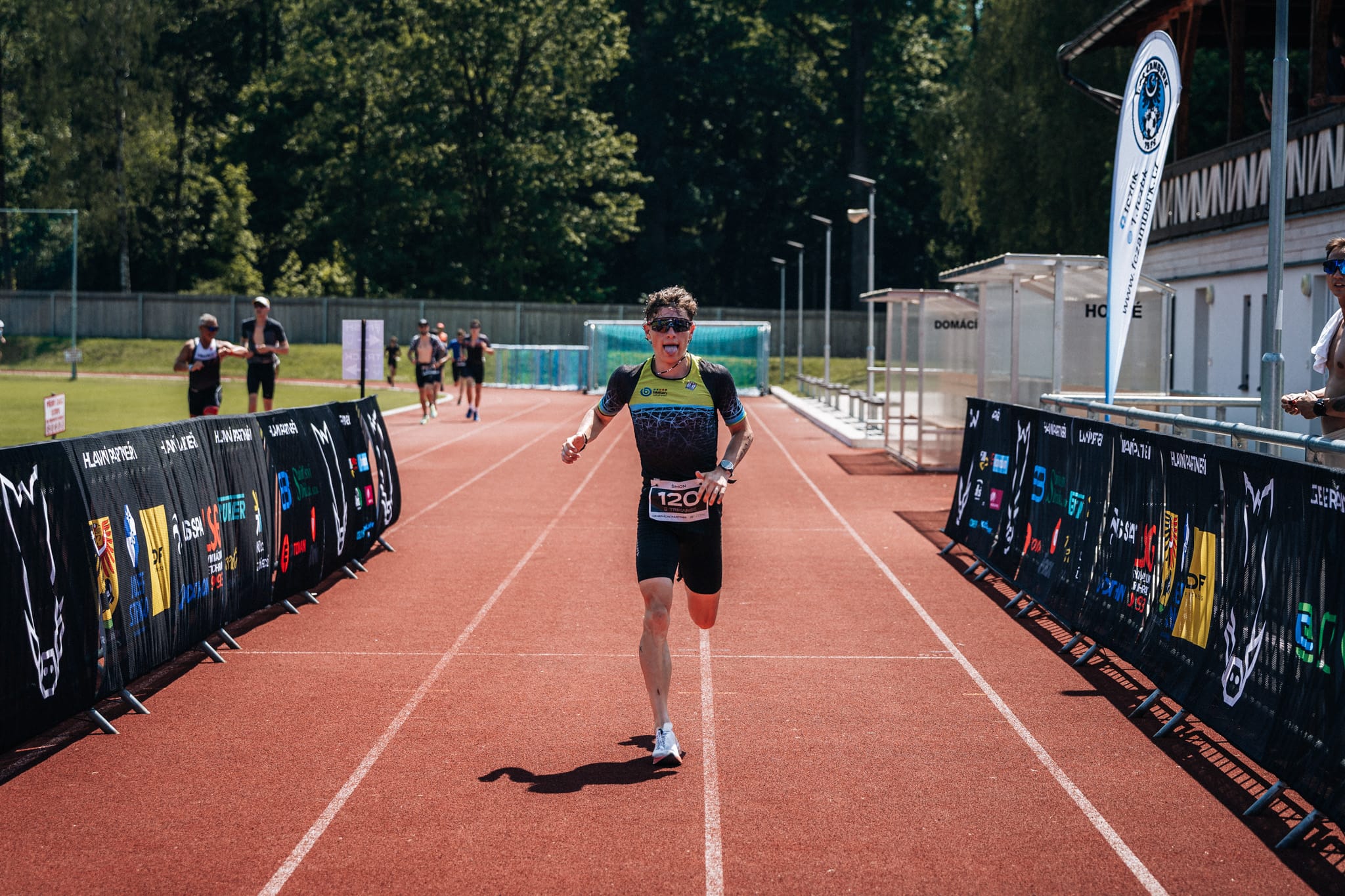 Vyčerpaný triatlonista s vyplazeným jazykem finišuje na rovince atletické dráhy