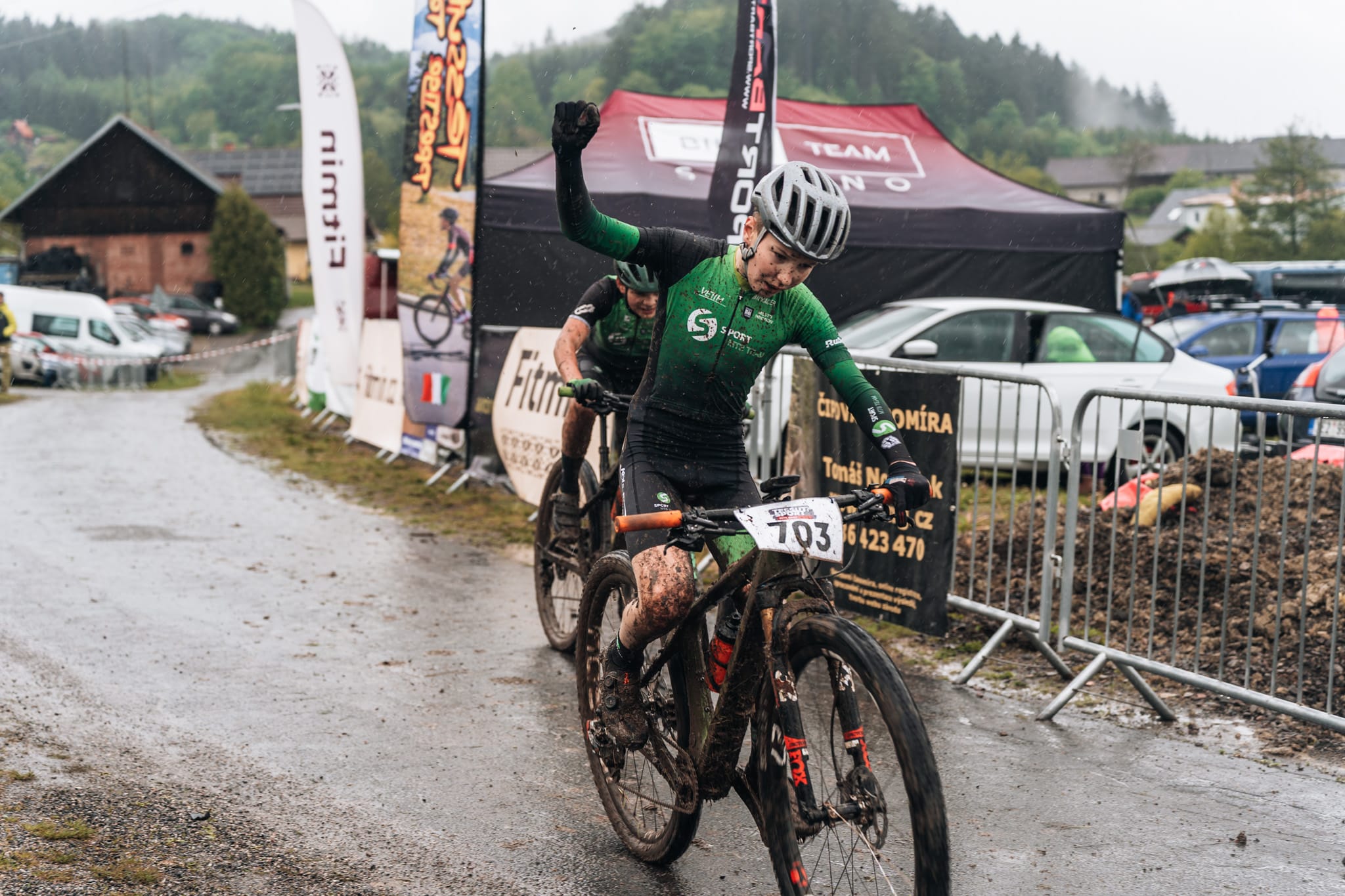 Zablácená bikerka v zeleném dresu zvedá pěst při dojezdu do cíle v dešti