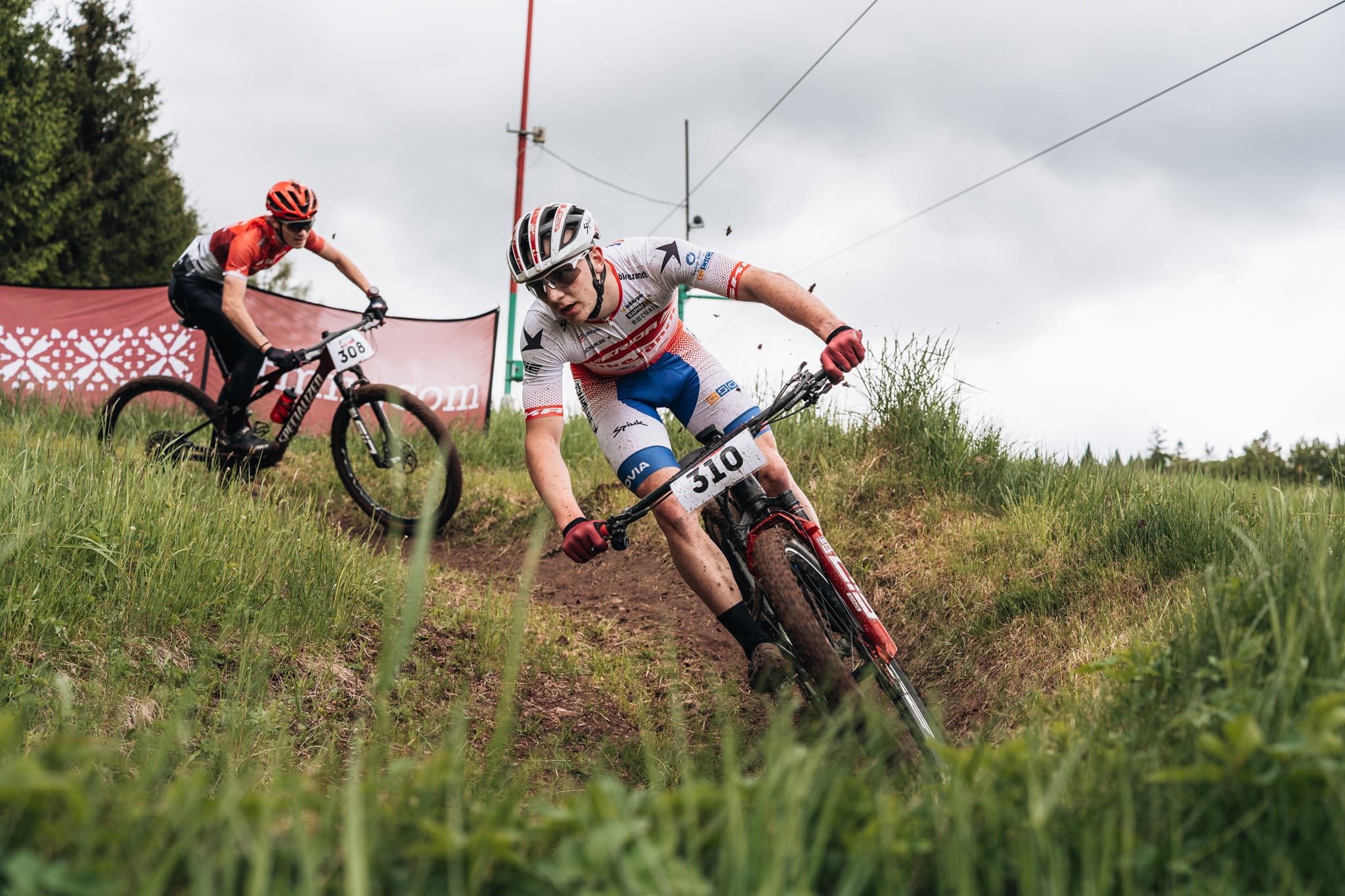 Dva bikeři se řítí travnatou zatáčkou nad sebou, první v bílo-červeném dresu