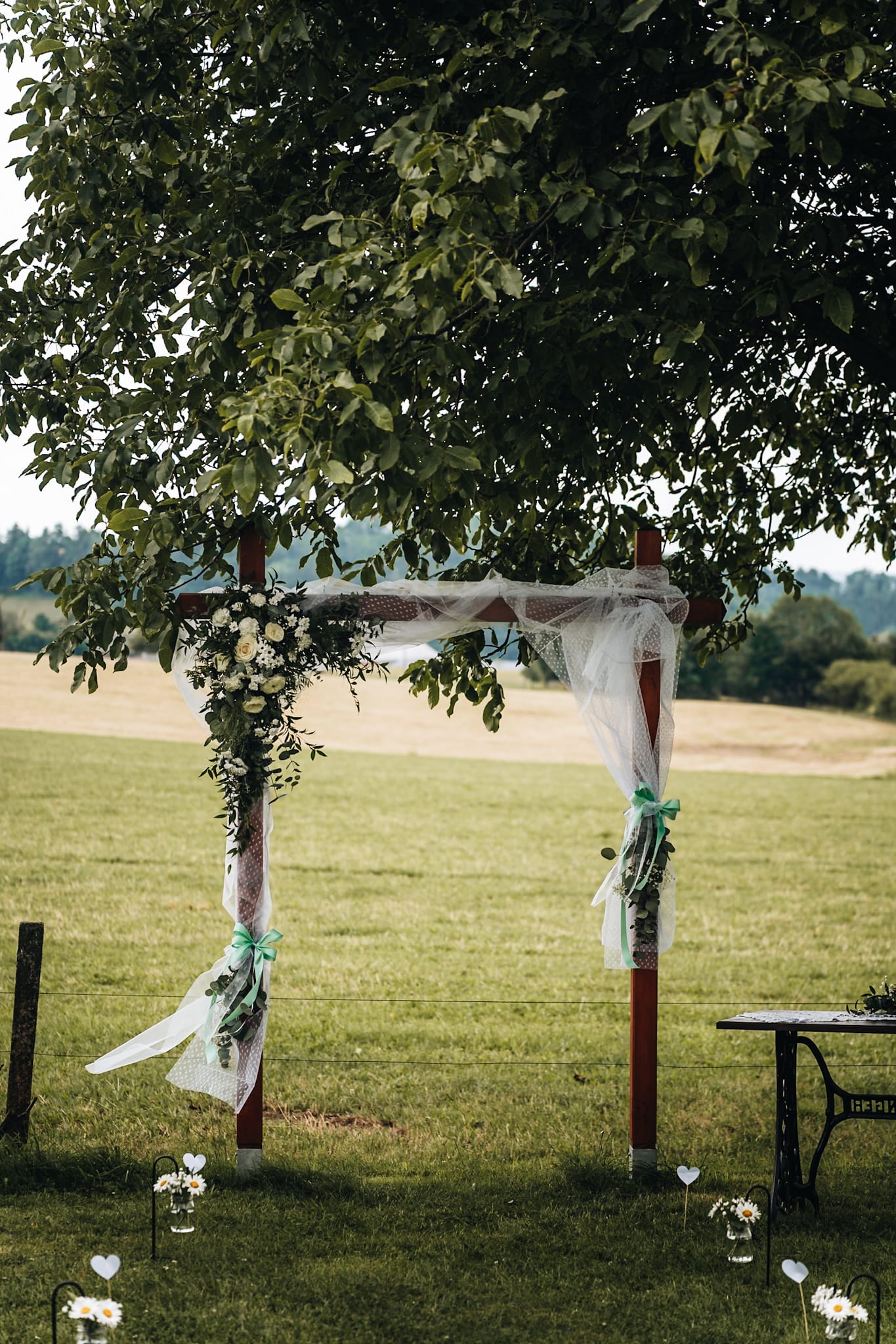 Dřevěná svatební brána zdobená bílými růžemi, tylem a mentolovými mašlemi pod korunou ořešáku